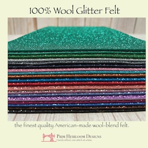 Op de afbeelding: Een stapel 100% wol glittervilt vellen in verschillende kleuren, waaronder groen, grijs, blauw, rood, roze, paars en bruin. Het vilt wordt beschreven als "het beste Amerikaanse wolmengsel vilt". De tekst "PRIM HEIRLOOM DESIGNS Handgemaakt, &eacute;&eacute;n steek tegelijk." is ook zichtbaar.