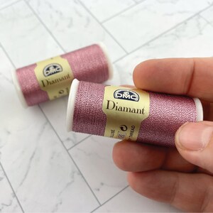 May include: Two spools of DMC Diamant thread in a shimmering rose color. One spool is held by a hand. The spools have white ends and a gold label with the DMC logo and the word "Diamant".