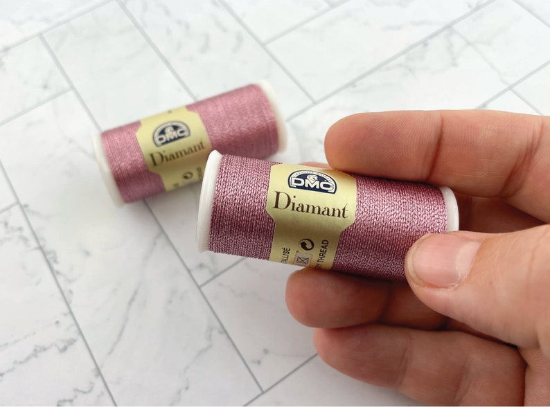May include: Two spools of DMC Diamant thread in a shimmering rose color. One spool is held by a hand. The spools have white ends and a gold label with the DMC logo and the word "Diamant".