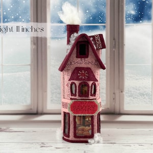 May include: A pink felt bakery building with a white chimney and a sign that reads "BAKERY". The building is 11 inches tall and has a window with a view of bread inside.