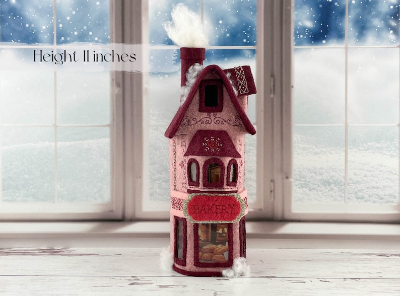 May include: A pink felt bakery building with a white chimney and a sign that reads "BAKERY". The building is 11 inches tall and has a window with a view of bread inside.
