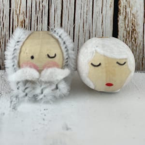 May include: Two wooden ornaments with painted faces. One ornament has a white beard and mustache, the other has white hair. Both have closed eyes and red lips.