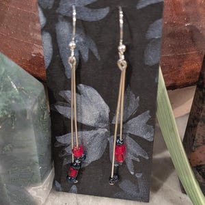 May include: A pair of silver dangle earrings with red and black beads. The earrings are hanging from a black card with a floral design.