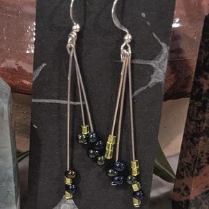 May include: A pair of silver dangle earrings with black and gold beads. The earrings are hanging from a black display card.