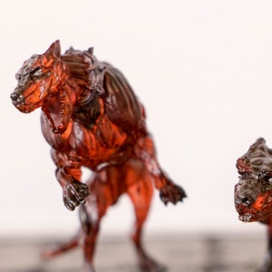 Może przedstawiać: Dwie miniaturowe figurki czerwonych, przezroczystych, trójgłowych psów z otwartymi paszczami, stojące na kamiennej powierzchni.