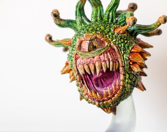 May include: A green and orange Beholder monster figurine with multiple eyes and a large open mouth with sharp teeth. The figurine is made of painted resin and is a popular collectible for fantasy game enthusiasts.
