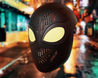 May include: A black mask with a textured finish and yellow eye-shaped details. The mask features a perforated pattern on the top and mouth area, similar to a superhero mask. The background shows a blurred city street.