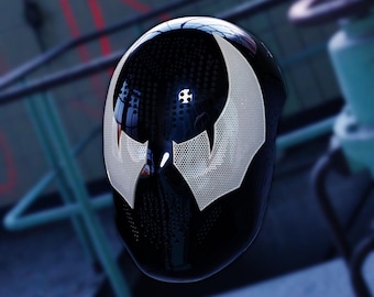 May include: A glossy black helmet with a white, perforated design on the face, resembling a stylized mask. The helmet reflects the surrounding environment, with a focus on the mask's unique shape and texture. The design is reminiscent of a comic book character.