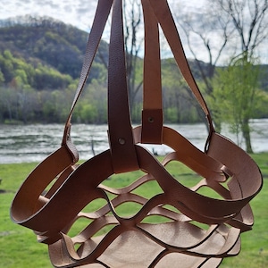 May include: A brown leather basket with a woven design hangs from a hook. The basket is made of thick leather strips that are woven together to create a lattice pattern. The basket is empty and has a rustic look.