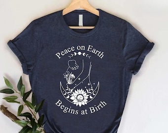 May include: A navy blue t-shirt with a white graphic design. The graphic features a crescent moon, a sunflower, and two hands holding a flower. The text "Peace on Earth Begins at Birth" is printed below the graphic.