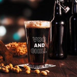 以下が含まれることがあります： 「VOO AND CHILL」の白い文字が入った、泡の立った濃い色のビールが入ったグラス。コースターの上に置かれ、背景にはピーナッツとボトルが配置されています。木製の表面に置かれたシーンです。