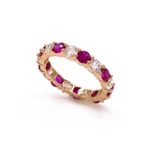 May include: A gold ring featuring alternating round-cut rubies and diamonds. The gemstones are set in a continuous pattern around the band. The ring is displayed on a white background, highlighting the sparkle of the gems.