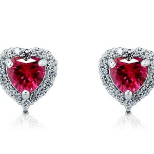 May include: A pair of silver heart-shaped earrings with a red heart-shaped gemstone in the center of each earring. The gemstones are surrounded by a halo of smaller clear gemstones.