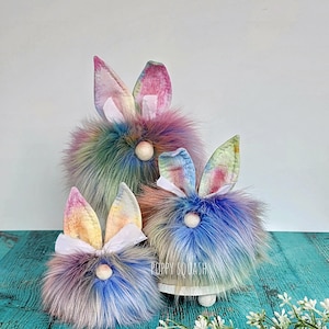 May include: Three colorful Easter bunny gnomes with tie-dye fabric ears and fluffy fur. The gnomes are arranged in a pyramid shape on a wooden surface.