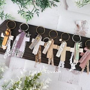 May include: A collection of handmade cross keychains with various embellishments, including lace, beads, and charms. Each keychain features a cross shape with unique textures and colors, attached to a metal keyring. The keychains are displayed on a dark wooden board.