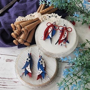 May include: Two pairs of dangle earrings with red, white, and blue fabric and silver star accents. The earrings are displayed on two white wooden stands.