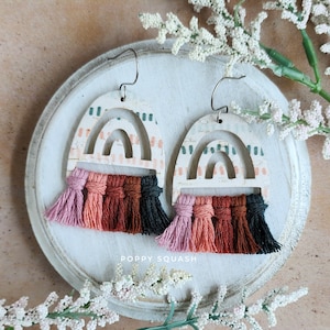 May include: A pair of white wooden earrings with a rainbow cutout design and colorful tassel fringe. The earrings are on a white background with the text "POPPY SQUASH" visible.