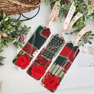 May include: Three handmade Christmas gift tags with red and green patchwork fabric, each with a red fabric rose and a white ribbon with red text that says "Merry Christmas" or "Merry".