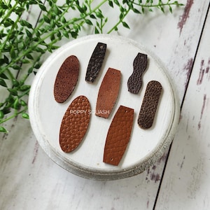 May include: Six leather pieces with holes for stitching, in various shapes and sizes. The pieces are brown, dark brown, and black. The pieces are arranged on a white surface with the text "POPPY SQUASH" visible.