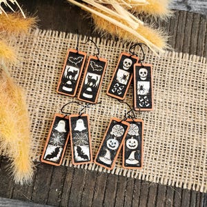 May include: A collection of Halloween-themed earrings. Each earring is a rectangular shape with a black background and orange border. The designs include cats, bats, ghosts, skulls, spiders, and jack-o'-lanterns. The earrings are hanging on black cords.