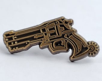 May include: A gold-colored metal pin shaped like a revolver. The pin has a detailed design and a textured surface.