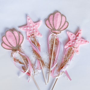 May include: Four pink and gold mermaid wands with white and pink ribbons. The wands are decorated with seashells and starfish.