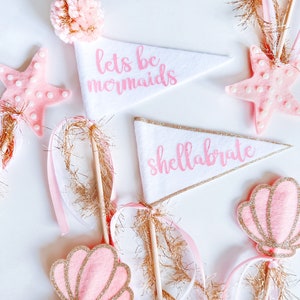 May include: A set of pink and gold mermaid themed party decorations. The decorations include a white felt pennant with the text "lets be mermaids", a white felt pennant with the text "shell-abrate", a pink and gold starfish, a pink and gold seashell, and a pink and gold pom-pom.