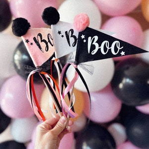 Könnte beinhalten: Drei Halloween-Wimpel mit dem Wort "Boo" in weißer Schrift. Die Wimpel sind rosa, weiß und schwarz, jeder mit einem Pompon und Sternen verziert. Bunte Bänder und Tüll sind an den Holzstäben befestigt.