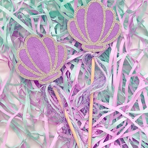 May include: Two purple glitter seashell wands with white and light blue tulle streamers. The wands are on a bed of pink and light blue shredded paper.