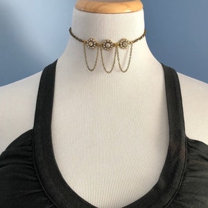 Puede incluir: Un collar gargantilla dorado con tres colgantes en forma de flor, cada uno con diamantes de imitación transparentes. El collar tiene cadenas colgantes con eslabones pequeños.