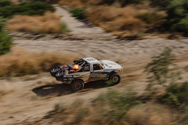 Puede incluir: Un cami&oacute;n todoterreno blanco con ruedas negras y un logotipo Eat My Wake negro y rojo en el lateral circula por un camino de tierra. El cami&oacute;n levanta polvo y tierra al conducir.