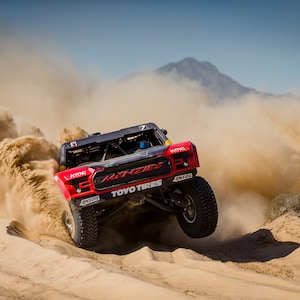 May include: A red race car with black and white accents drives through a desert landscape, kicking up a cloud of dust. The car has "TOYO TIRES" and "MENZIES" written on the side.