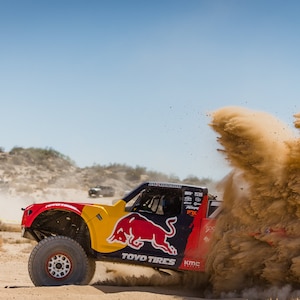 May include: A red, yellow, and black off-road truck with a Red Bull logo drives through a cloud of brown dust. The truck has "TOYO TIRES" and "KMC" written on it.
