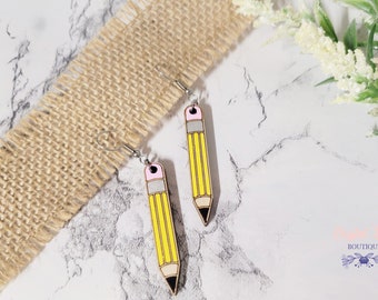 May include: A pair of yellow pencil earrings with pink erasers and black lead. The earrings are made of wood and have silver hooks.