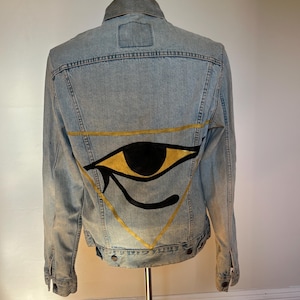 May include: Light blue denim jacket with a gold and black eye design on the back. The eye is within a gold triangle. The jacket has a gray collar and a classic button-up design.