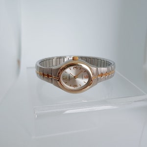 Puede incluir: Un reloj de pulsera analógico plateado y dorado con una esfera blanca y marcadores de hora en números romanos negros. El reloj tiene una pulsera de metal plateado.