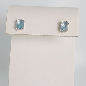 Peut inclure: Une paire de petites boucles d'oreilles en pierres précieuses bleu clair avec un sertissage argenté. Les boucles d'oreilles sont présentées sur une carte blanche.