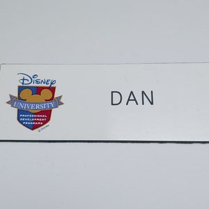 May include: White name tag with a black border. The left side features the Disney University logo with a red, yellow, and blue color scheme. The right side has the name "DAN" printed in black.