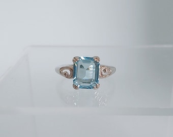May include: A silver ring featuring a light blue square-shaped gemstone and delicate swirls on the band.