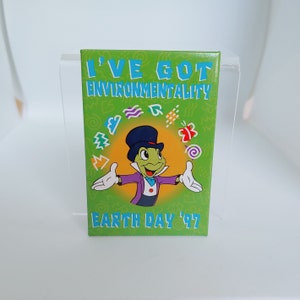 Peut inclure: Bouton vert et jaune avec un personnage de dessin animé de Jiminy Cricket de Pinocchio de Disney. Le bouton indique "I've Got Environmentality Earth Day '97".
