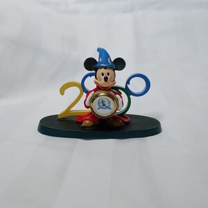 May include: A Mickey Mouse figurine wearing a blue sorcerer's hat, holding a gold clock with the Walt Disney World logo. The figurine is standing on a black base with the numbers 2000 and the iconic Mickey Mouse ears.