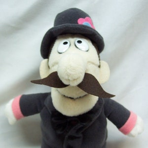 May include: A plush doll of a man in a dark gray suit, black shoes, and a black hat with a pink and blue flower. The doll has a white face, large eyes, a brown mustache, and a black bow tie.
