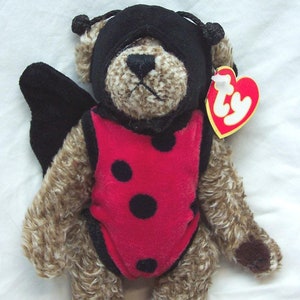 May include: A brown and white fuzzy teddy bear wearing a black ladybug costume with red and black polka dots and a black eye mask. The bear is holding a red Ty tag with the word "Ty" in white.