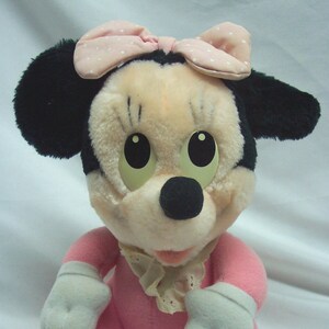 May include: A pink plush toy of Mickey Mouse, wearing a pink and white polka dot bow and a white lace collar. The toy has black ears and a black nose.