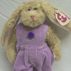 May include: A plush tan rabbit stuffed animal with long ears, wearing lavender corduroy overalls featuring a small embroidered purple flower on the chest. A heart-shaped Ty Collectible tag is attached to the left ear, displaying the Ty logo.