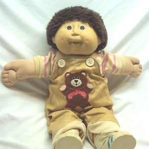 Op de afbeelding: Een Cabbage Patch Kid pop met bruin haar, die een bruine overall draagt met een bruine teddybeer-patch en een gestreept shirt.