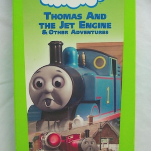 Puede incluir: Una cinta VHS de "Thomas & Friends: Thomas and the Jet Engine & Other Adventures". La portada presenta un tren azul con el número 1 y dos trenes rojos. El título está en una pancarta blanca en forma de nube.