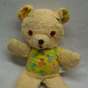 Può includere: Un orsacchiotto vintage con corpo color crema, occhi marroni e naso nero. L'orso indossa un completo giallo con forme geometriche colorate. Le orecchie e i piedi hanno un tessuto fantasia abbinato.