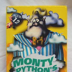 May include: A yellow VHS tape case with a cartoon illustration of a man with a large nose and a striped shirt. The text "Monty Python's Flying Circus" is written in green and white letters. The case also features a blue pot with steam coming out of it and the text "Contains: Job Hunter, Spam & Cannibalism".