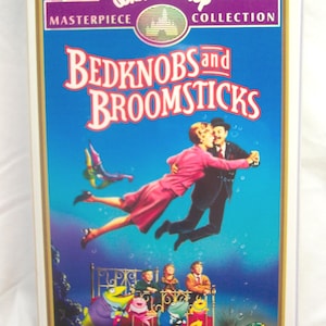 May include: VHS tape for the Disney animated film "Bedknobs and Broomsticks" from the Masterpiece Collection. The cover features a couple dancing in the air above a bed with animated frogs and children. The background is a vibrant blue with the film's title.